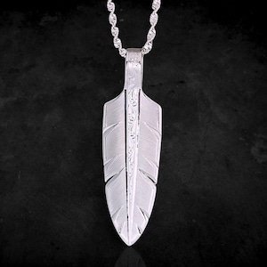 May include: A silver-toned pendant shaped like a feather, featuring intricate engraved scrollwork along the central spine and a brushed metal texture on the vanes. The pendant hangs from a matching silver-toned twisted rope chain necklace.
