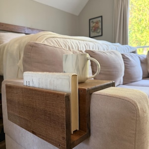 Puede incluir: Una mesa de brazo de sofá de madera con un sujetalibros incorporado. Una taza de mármol blanco y un libro están sobre la mesa. La mesa es de madera marrón oscuro y está unida a un sofá rosa claro.