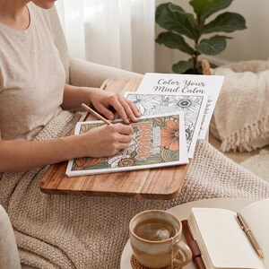 May include: A person coloring in a floral-themed coloring book with the words "Color Your Mind Calm". A wooden tray holds the book and a pencil. A cup of tea and a notebook are on a side table.