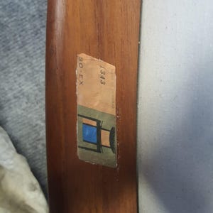 May include: Close-up of a wooden chair arm with a sticker. The sticker has the text "BO-EX" and "1343 Made in Denmark" along with a small illustration of a chair with a blue seat. The wood has a warm, brown tone.