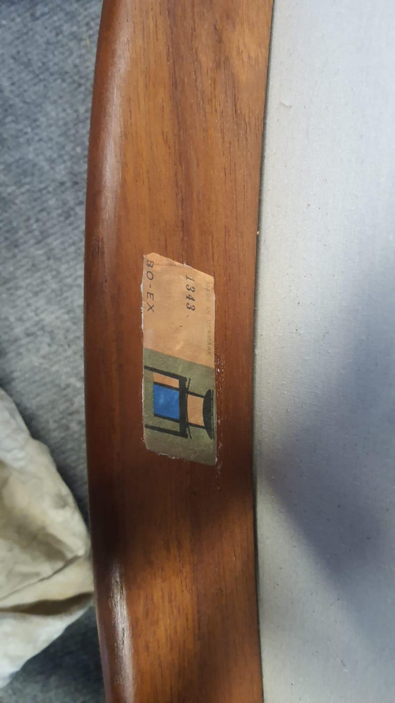May include: Close-up of a wooden chair arm with a sticker. The sticker has the text "BO-EX" and "1343 Made in Denmark" along with a small illustration of a chair with a blue seat. The wood has a warm, brown tone.