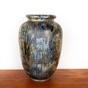 Peut inclure: Un grand vase en céramique avec un corps arrondi et un bord évasé. Le vase présente un glaçage moucheté dans des tons de bleu, de brun et de vert, créant un aspect texturé. Il est posé sur une surface en bois sur fond blanc.