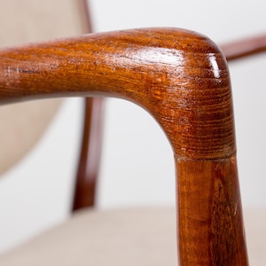 May include: Close-up of a wooden armchair armrest. The armrest is made of polished, reddish-brown wood with a curved design. The chair has a light-colored fabric seat and back.
