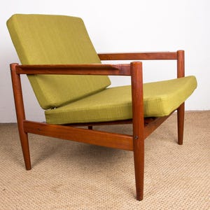 Peut inclure: Fauteuil de style milieu du siècle avec une structure en bois et des coussins vert olive. La chaise a un profil bas avec des pieds et des accoudoirs inclinés. Les coussins semblent être faits d'un tissu texturé. La chaise est placée sur un fond neutre.