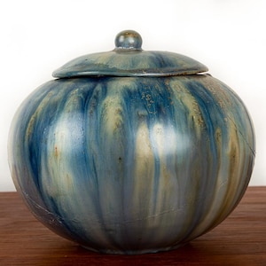 Peut inclure: Un pot en céramique avec un couvercle, de forme ronde et avec un glaçage bleu et beige. Le pot a un petit bouton sur le couvercle. Le glaçage présente un motif vertical et fluide.