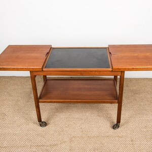 May include: A mid-century modern wooden bar cart with a rectangular black center surface and two extending side panels. The cart has a lower shelf and rolls on small caster wheels. The wood has a warm, reddish-brown tone.