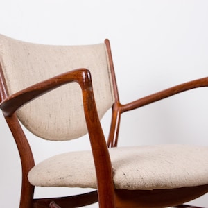 May include: A mid-century modern armchair with a curved wooden frame and light-colored fabric upholstery. The chair features a rounded seat and backrest, and curved armrests. The wood has a rich, dark finish, and the chair is set against a white background.