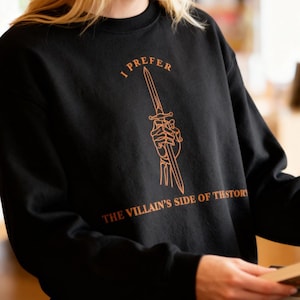 K&ouml;nnte beinhalten: Schwarzes Sweatshirt mit dem Text "I PREFER THE VILLAIN'S SIDE OF HISTORY" in Orange. Das Design zeigt einen Dolch, der von einer Skeletthand gehalten wird. Das Sweatshirt wird von einer Person in einer Buchhandlung getragen.