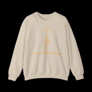 K&ouml;nnte beinhalten: Cremefarbenes Sweatshirt mit langen &Auml;rmeln. Auf der Vorderseite befindet sich eine orangefarbene Illustration einer Skeletthand, die ein Schwert h&auml;lt, mit dem Text "I PREFER THE VILLAIN'S SIDE OF THE STORY" in Orange.