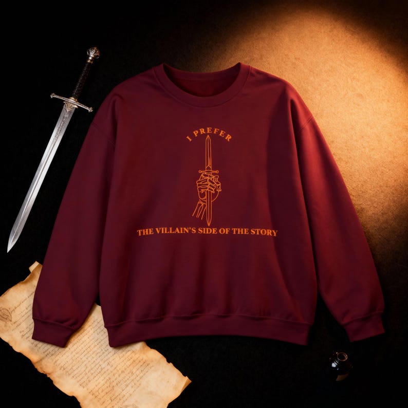 K&ouml;nnte beinhalten: Burgunderfarbenes Sweatshirt mit dem orangefarbenen Text "I Prefer the Villain's Side of the Story". Das Design zeigt eine Hand, die ein Schwert h&auml;lt. Im Hintergrund ist ein silbernes Schwert zu sehen.