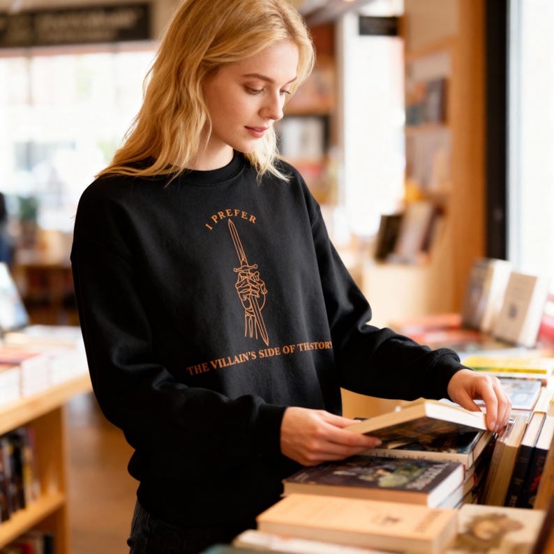 K&ouml;nnte beinhalten: Schwarzes Sweatshirt mit dem Text "I PREFER THE VILLAIN'S SIDE OF HISTORY" in Orange. Das Design zeigt einen Dolch, der von einer Skeletthand gehalten wird. Das Sweatshirt wird von einer Person in einer Buchhandlung getragen.