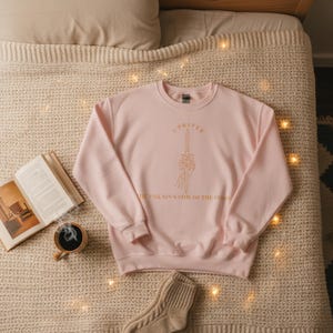 K&ouml;nnte beinhalten: Hellrosa Sweatshirt mit goldenem Text "I PREFER THE VILLAIN'S SIDE OF THE STORY" und einem Schwert, das von einer Skeletthand gehalten wird. Das Sweatshirt liegt auf einer beigefarbenen Strickdecke.