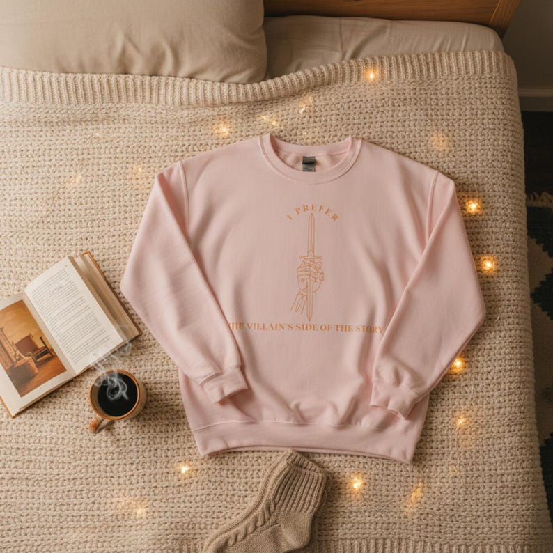 K&ouml;nnte beinhalten: Hellrosa Sweatshirt mit goldenem Text "I PREFER THE VILLAIN'S SIDE OF THE STORY" und einem Schwert, das von einer Skeletthand gehalten wird. Das Sweatshirt liegt auf einer beigefarbenen Strickdecke.