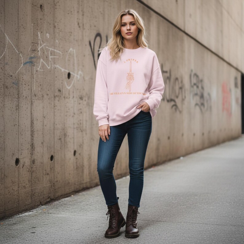 K&ouml;nnte beinhalten: Hellrosa Sweatshirt mit dem Text "I Prefer The Villain's Side of the Story" in Kupferfarbe. Das Sweatshirt hat eine Grafik von einem Schwert und einer Hand. Die Person tr&auml;gt eine dunkelblaue Jeans und braune Stiefel.