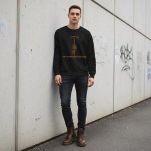 K&ouml;nnte beinhalten: Schwarzes Sweatshirt mit goldenem Text "I Prefer The Villain's Side of the Story" und einer Grafik einer Skeletthand, die ein Schwert h&auml;lt. Das Design befindet sich mittig auf der Brust.
