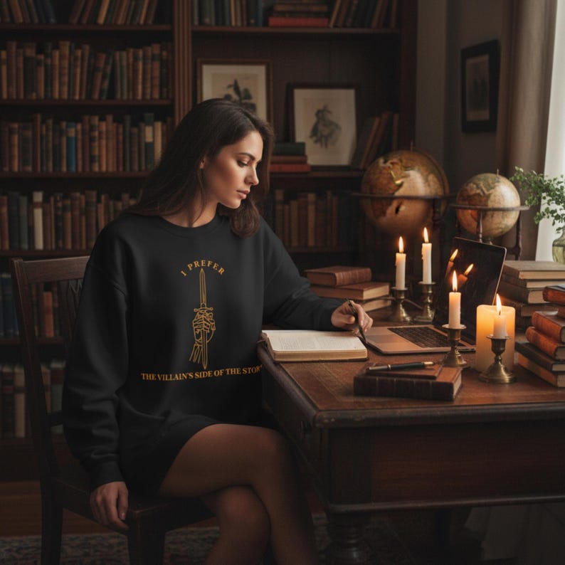 K&ouml;nnte beinhalten: Schwarzes Sweatshirt mit goldenem Text "I Prefer The Villain's Side of the Story" und einer Grafik eines Schwertes, das von einer Skeletthand gehalten wird. Die Person tr&auml;gt das Sweatshirt und sitzt an einem Holztisch.