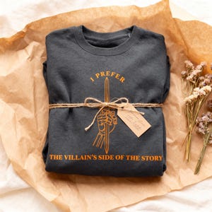 K&ouml;nnte beinhalten: Dunkelgraues Sweatshirt mit dem Text "I PREFER THE VILLAIN'S SIDE OF THE STORY" in Orange. Das Design zeigt ein Schwert, das von einer Hand gehalten wird. Das Sweatshirt ist mit Bindfaden und einem Etikett verpackt.