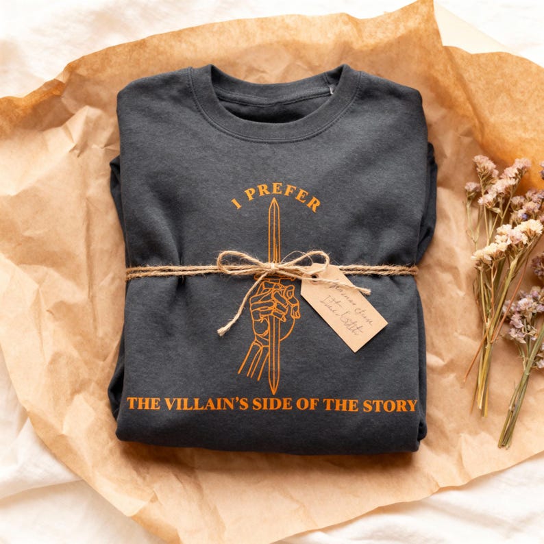 K&ouml;nnte beinhalten: Dunkelgraues Sweatshirt mit dem Text "I PREFER THE VILLAIN'S SIDE OF THE STORY" in Orange. Das Design zeigt ein Schwert, das von einer Hand gehalten wird. Das Sweatshirt ist mit Bindfaden und einem Etikett verpackt.