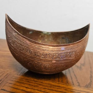 May include: A crescent-shaped copper bowl featuring an intricate embossed pattern of swirling vines and geometric lines along the exterior. The metal has a warm, aged patina with visible variations in color, and the rim curves upward at both ends.