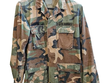 Puede incluir: Chaqueta con estampado de camuflaje con múltiples bolsillos delanteros y el texto "U.S. ARMY" encima de un bolsillo. La chaqueta es en tonos verdes, marrones y negros. Cuelga de una percha negra.