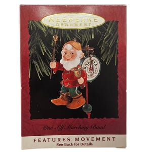 May include: Keepsake Ornament featuring a cheerful elf in a red coat, green pants, and brown boots, holding a baton and a drum. The ornament is set against a black background with a Christmas tree branch. Text on the box reads "One-Elf Marching Band."