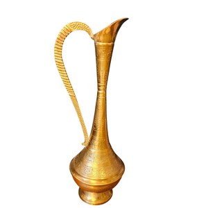 May include: A tall, slender brass pitcher featuring a long, narrow neck and a wide, rounded base. The surface is decorated with intricate, etched floral and geometric patterns. It includes a curved, textured handle and a pointed pouring spout.