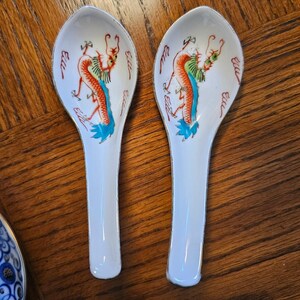 May include: Two white ceramic soup spoons featuring a hand-painted dragon motif in orange, green, and blue. Each spoon has a long, slender handle and a deep, rounded bowl with a pointed tip, accented by a thin silver-colored rim along the edges.