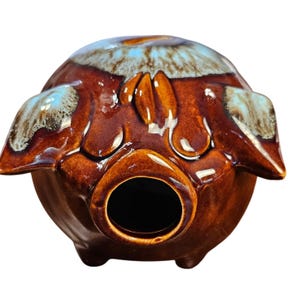 May include: A ceramic pig-shaped bank with a glossy brown glaze. The pig has a round body, small ears, and a snout with a large opening. The top of the pig is decorated with blue and white accents. The bank sits on three small feet.