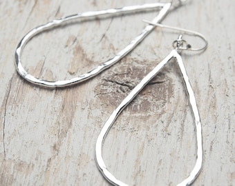 May include: A pair of silver teardrop earrings with a hammered texture. The earrings have a simple, elegant design with a hook closure. The earrings are displayed on a light-colored wooden surface.