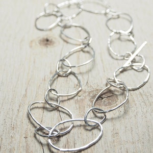May include: A silver necklace made of interconnected oval links. The necklace is laid out on a light wood surface.