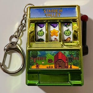 Puede incluir: Un llavero de máquina tragamonedas Stardew Valley verde y dorado. La parte superior del llavero dice "Stardew Valley". La máquina tragamonedas muestra varios iconos del juego. Un mango rojo está en el lateral y una cadena y un cierre plateados están adjuntos.