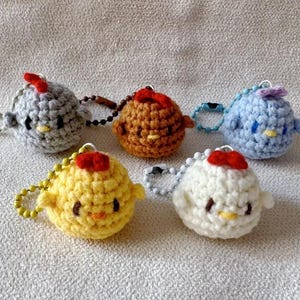 Puede incluir: Colección de cinco llaveros de pollitos de ganchillo en varios colores: gris, marrón, azul, amarillo y blanco. Cada pollito tiene un lazo rojo, ojos negros pequeños y un pico amarillo. Los llaveros están sujetos a anillas metálicas.