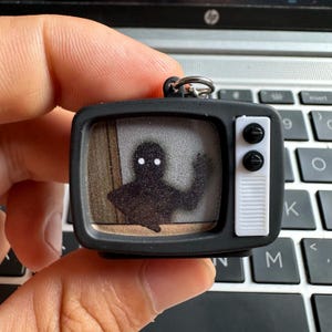 Può includere: Un portachiavi a forma di televisore nero in miniatura con una figura silhouette sullo schermo. La figura ha occhi bianchi luminosi e sta salutando. Il televisore ha pulsanti bianchi e un portachiavi argentato.