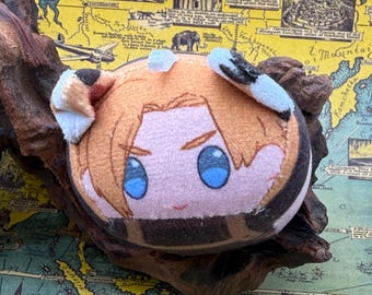 May include: A round plush toy with a cartoon character face, featuring blue eyes, light brown hair, and tan ears with black tips. The toy is resting on a piece of dark wood, with a vintage map in the background.