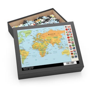 Puede incluir: Una caja negra que contiene un rompecabezas de mapa del mundo. El rompecabezas muestra un mapa detallado con países etiquetados en varios colores. La caja también contiene piezas sueltas del rompecabezas. La caja dice "16x20 Puzzle 500 pcs Ages 9+" y tiene un código de barras.