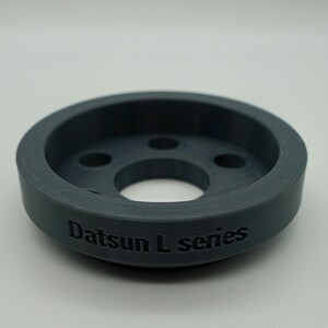Op de afbeelding: Een donkergrijze, cirkelvormige auto-onderdeel met een centraal gat en verschillende kleinere gaten. De tekst "Datsun L series" is in reliëf op de zijkant aangebracht. Het onderdeel heeft een verhoogde buitenrand en een verzonken binnenruimte.