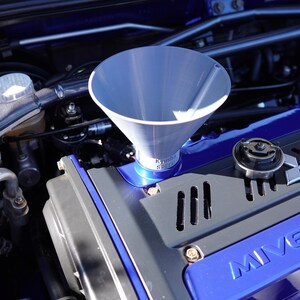 Puede incluir: Un embudo plateado se coloca en el motor de un coche azul. El motor tiene componentes negros y azules, incluyendo el texto "MIVEC TURBO". La imagen está tomada a la luz del día.