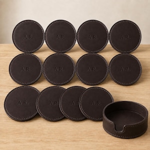 May include: A set of ten circular dark brown coasters, each featuring the embossed initials A.R. in the center. The coasters are arranged in three rows, accompanied by a matching dark brown cylindrical holder with a cutout side detail.