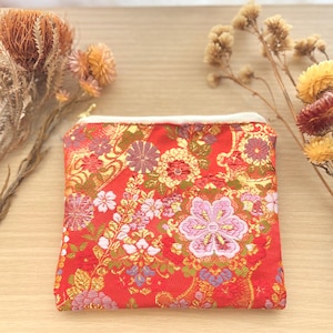 May include: A small, square red pouch with a white zipper and a floral pattern in shades of pink, gold, and purple. The pouch is made of a textured fabric and is likely used for carrying small items. Dried flowers are in the background.