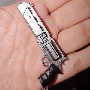 May include: A miniature silver-colored metal revolver keychain featuring a long barrel with rectangular cutouts, a rotating cylinder, a curved trigger guard, and a black ergonomic grip handle with textured detailing on the side.