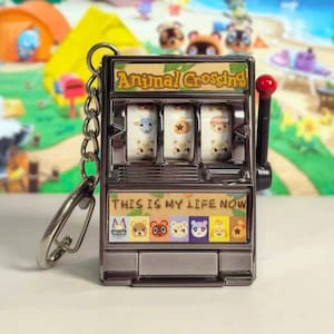 Op de afbeelding: Een zilverkleurige Animal Crossing gokautomaat sleutelhanger. De gokautomaat heeft drie rollen met karakterafbeeldingen en de tekst "Animal Crossing" bovenaan. De onderkant van de sleutelhanger leest "THIS IS MY LIFE NOW".