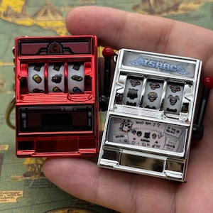 Może przedstawiać: Dwa miniaturowe breloki do kluczy w kształcie automatów do gier. Jeden jest czerwony, drugi srebrny. Czerwona maszyna ma białe bębny z różnymi symbolami. Srebrna maszyna ma napis "THE BINDING OF ISAAC" i "WHO AM I?" z przodu.