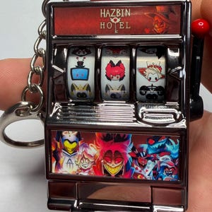 Könnte beinhalten: Ein silberner Schlüsselanhänger in Form eines Spielautomaten mit Charakteren aus Hazbin Hotel. Auf der Oberseite der Maschine steht "HAZBIN HOTEL" in roter Schrift. Die Walzen zeigen Cartoon-Charaktere. Auf der rechten Seite befindet sich ein roter Griff.