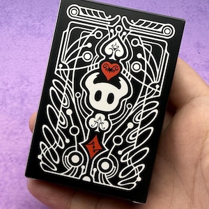 Può includere: Una scatola nera di carte da gioco con un design grafico bianco e rosso. Il design presenta un teschio stilizzato, cuori e altri elementi decorativi. La scatola è tenuta in mano su uno sfondo viola.