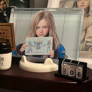 May include: A display featuring a framed image of a girl holding a drawing, a white ceramic cat-shaped object, a small black calendar displaying "DEC 06", and a coffee cup. The drawing depicts stick figures. The background includes a coffee menu.