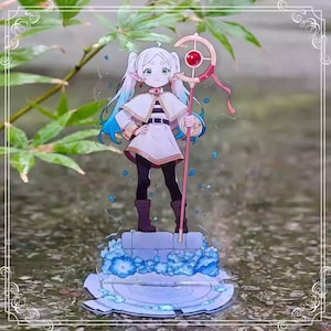 May include: A clear acrylic anime figure of a character with white hair, blue highlights, and pointed ears. The character wears a white outfit with a cape and holds a pink staff with a red orb. The figure stands on a base with blue floral accents.