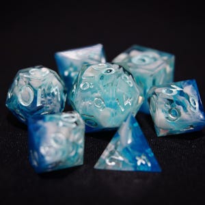 以下が含まれることがあります： 青と白のマーブル模様の7つの多面体サイコロのセット。各面に数字が見える、テーブルゲーム用のサイコロです。サイコロは黒い面に配置されています。