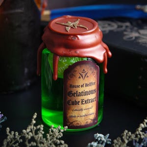 May include: A small, green glass bottle filled with a glowing green liquid, sealed with a red wax top. The label reads "House of Hellfire Gelatinous Cube Extract". The bottle is surrounded by dried plants and a dark, leather-bound book.