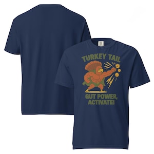 May include: Navy blue t-shirt featuring a cartoon turkey superhero with the text "TURKEY TAIL, GUT POWER, ACTIVATE!". The turkey is flexing its muscles. The shirt is a short-sleeved crew neck.