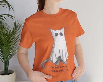 May include: Orange t-shirt featuring a cat dressed as a ghost. The cat is drawn in pencil with a white sheet and black eyes. The text "Spooky season feral edition" is below the cat.